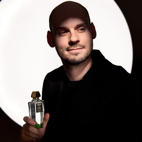 A man in a black jacket holding a fragrance bottle, illuminated by dramatic lighting.