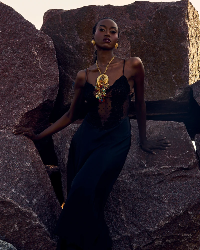 Model in a black gown with bold jewelry posing against stone backdrop.