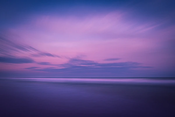 A serene sunset over calm waters, featuring soft purple and pink hues in the sky with gentle cloud movement.