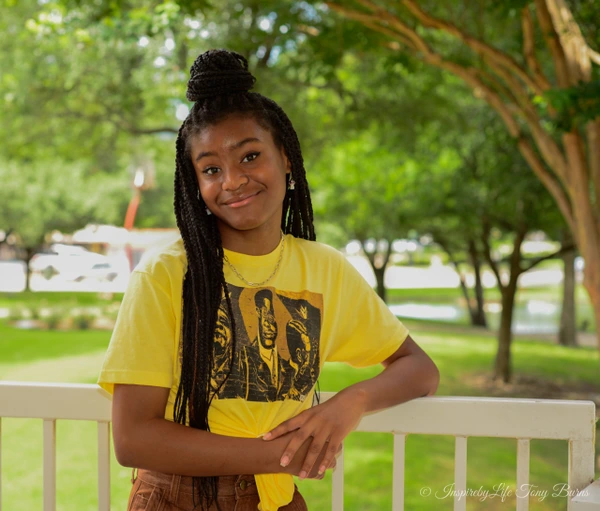 A young girl with braids smiles warmly while leaning on a railing in a green outdoor setting.