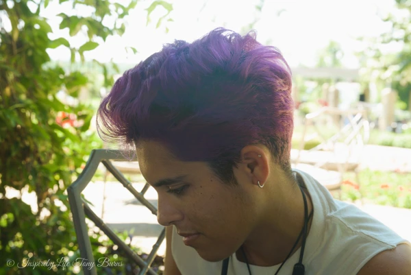 A young individual with purple hair sitting outdoors in a relaxed pose.