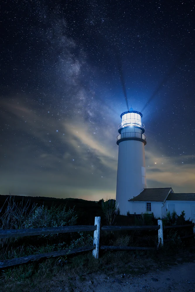 A lighthouse shines brightly under a star-filled Milky Way sky at night.