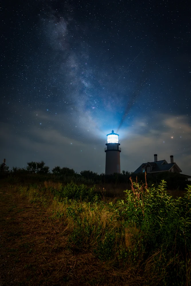 A lighthouse beams light under a star-filled sky with the Milky Way visible.