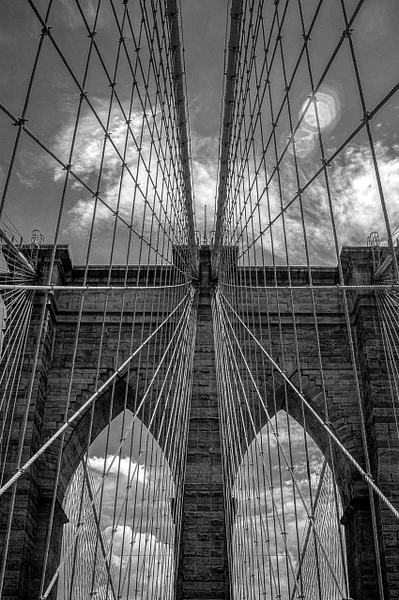Black-and-white upward view of Brooklyn Bridge cables converging around its stone tower beneath textured clouds