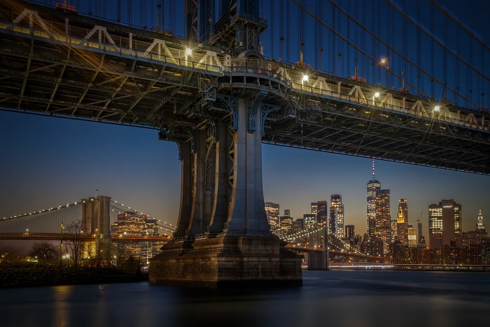 Illuminated Manhattan Bridge with city skyline at dusk reflecting on water.