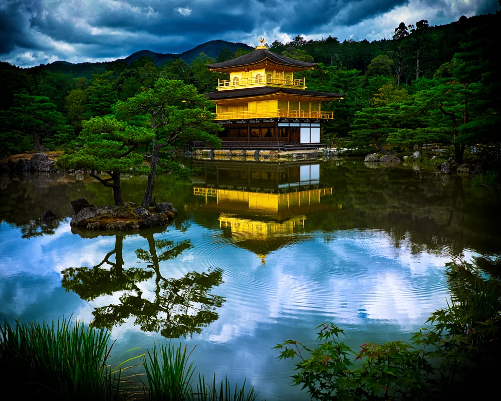 Kinkakuji Golden Pavilion reflecting in calm waters under dramatic clouds.
