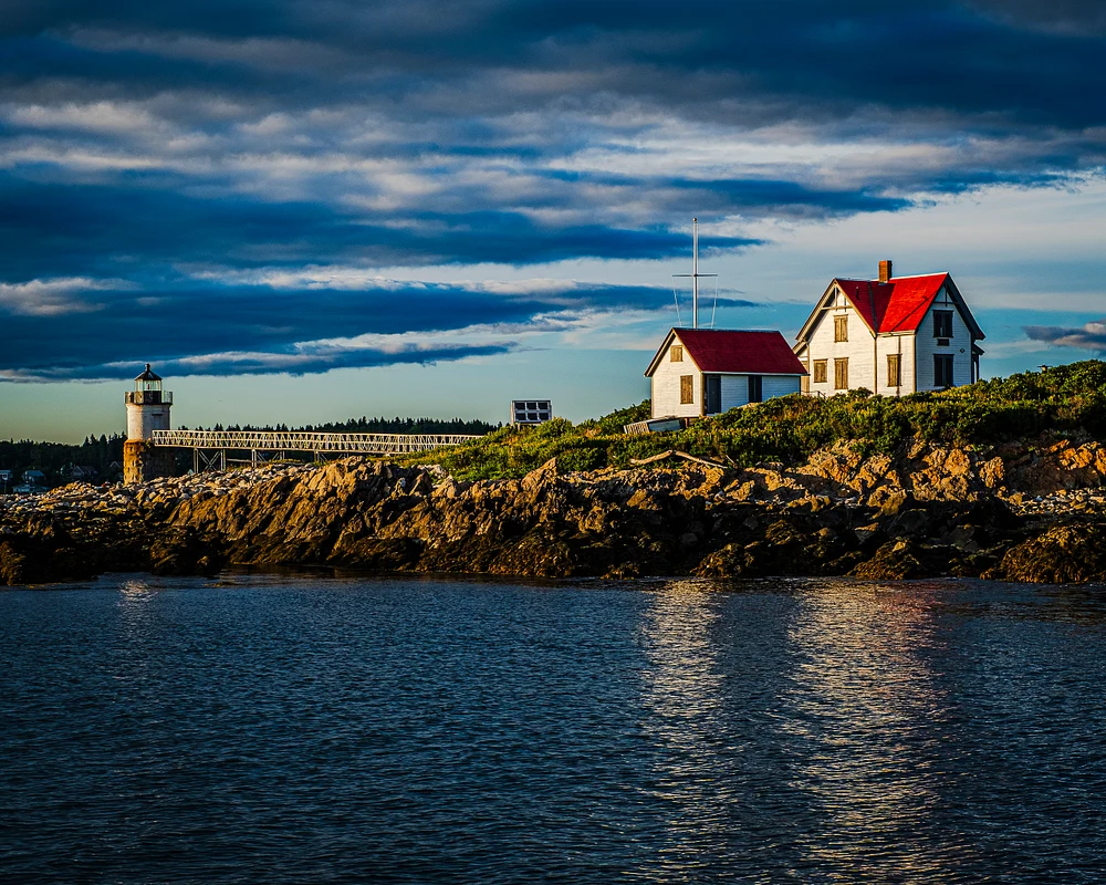 Cape Cod lighthouse and red-roofed house at dusk reflecting in calm waters.
