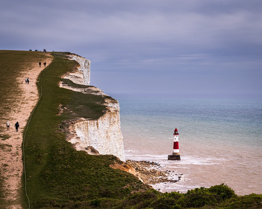 Beachy Head Lighthouse with chalk cliffs and visitors walking along the path.