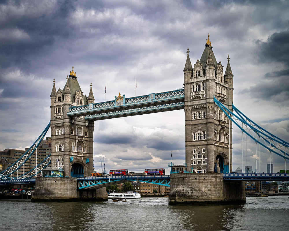 Tower Bridge in London with red buses crossing over the River Thames under a cloudy sky.