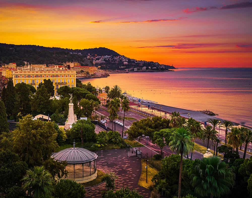 Sunset over Nice and the Promenade des Anglais, with palm trees, gardens, hillside homes, and calm sea