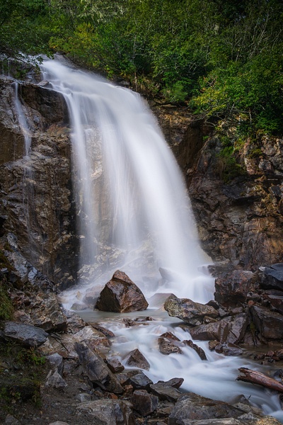 A commanding vertical fine art crop of the Mile 10 upper tier of Bridal Veil Falls along the Klondike Highway in Alaska. The composition tightly compresses the frame, showcasing a smooth, 2.5-second long-exposure water column that looks like a radiant white architectural pillar. Imposing, dark gray granite canyon walls tightly enclose the waterfall, channeling its downward rush into a deep, shadowed gorge.