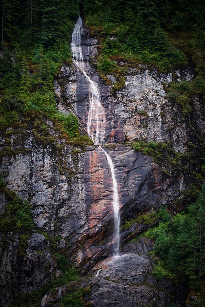 A high-contrast vertical fine art photograph of a narrow, ribbon-like waterfall cascading down a towering, dark gray granite cliff face in Endicott Arm fjord, Alaska. The wet, textured metamorphic rock is split vertically down the center by distinct, warm copper-red and iron mineral streaks. Deep shadows frame the left side, while lush green Pacific Northwest foliage clings to the mountain crevices, contrasting the bright white stream of water.