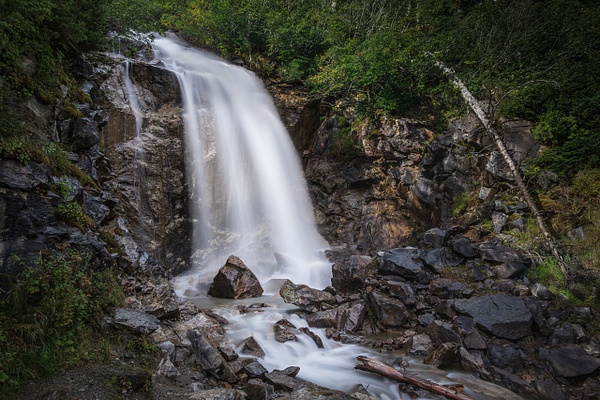 A horizontal long-exposure photograph capturing the upper sub-alpine tier of Bridal Veil Falls at Mile 10 of the Klondike Highway in Alaska. The 2.5-second exposure silences the water into a flawlessly smooth, mirror-like curtain of white glass. The luminous waterfall is bordered by sharp, rugged granite canyon walls, with subtle vignettes softly shading the top tree canopy and the swirling pool runoff at the bottom.