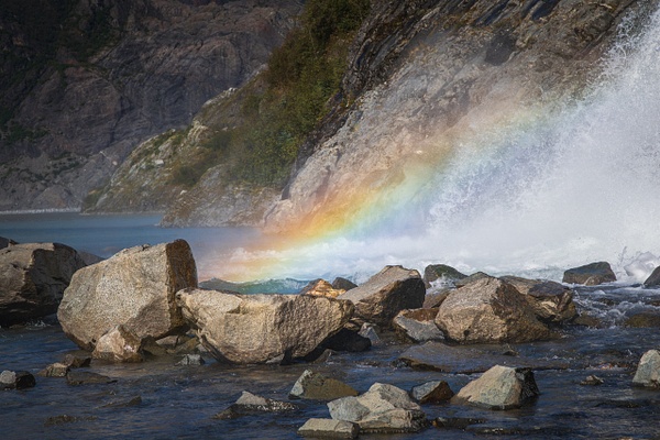 A vibrant, close-up fine art photograph captured at a fast 1/800th of a second shutter speed at the base of Nugget Falls in Juneau, Alaska. Millions of sharp, kinetic water droplets are frozen mid-air, refracting the late afternoon light into a brilliant, glowing rainbow arc. The colorful spectrum spans across massive, wet river boulders that showcase a warm, weathered golden-tan and iron-rich mineral texture.