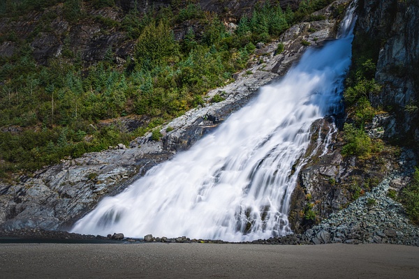 A sweeping, wide-angle horizontal landscape photograph of Nugget Falls dropping 377 feet down a massive, dark granite mountain cliff in Juneau, Alaska. The broad white waterfall empties directly onto a vast, flat glacial gravel bar on the edge of Mendenhall Lake. The entire scene holds a crisp, cool, high-contrast atmosphere, with a soft, uniform blanket of fine spray diffusing the river stones at the base of the cascade.