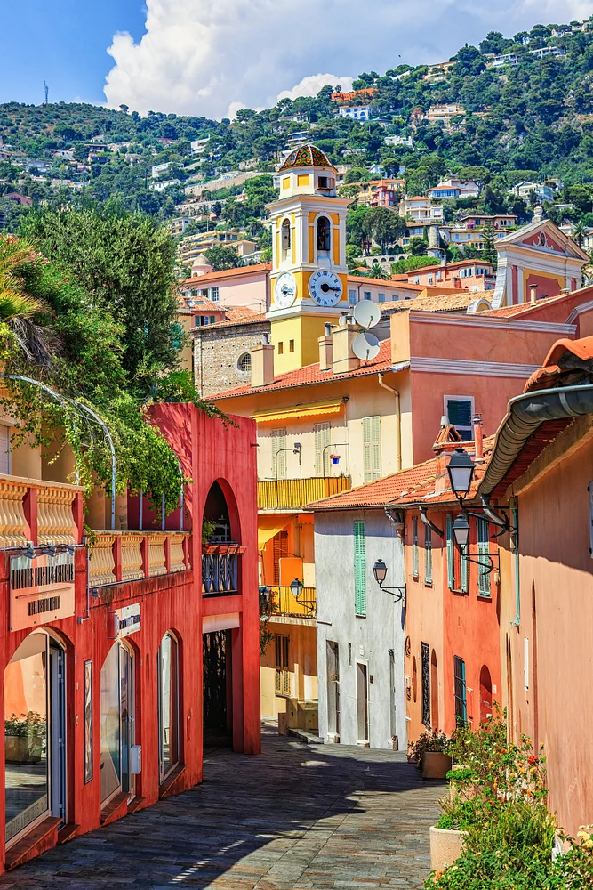 Colorful Villefranche-sur-Mer lane leading toward Saint-Michel clock tower, with green hills and terracotta roofs