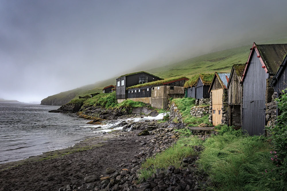 Misty Bour shoreline with traditional turf-roofed boathouses beside dark stones and calm North Atlantic water