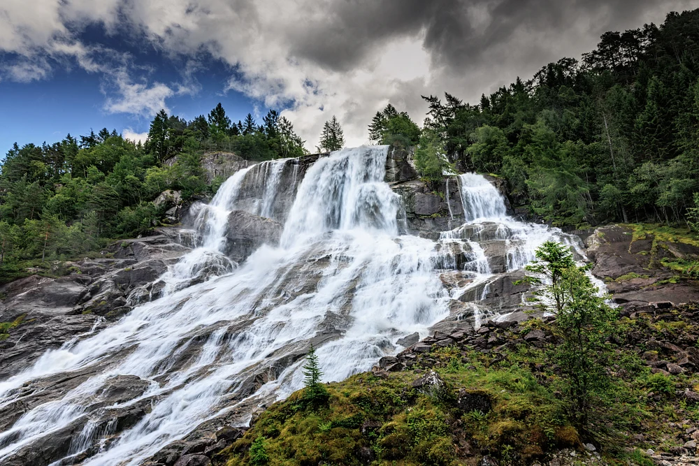 Furubergfossen waterfall spreads over dark rocks beneath forest, moss, and dramatic clouds near Odda, Norway