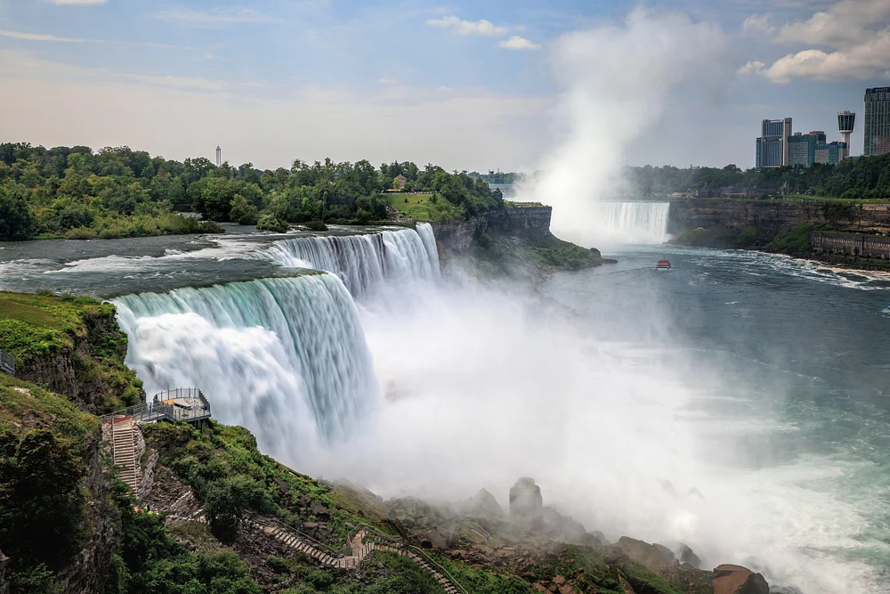 Wide view of Niagara Falls pouring into a misty gorge, with wooded banks, river rapids, and distant towers