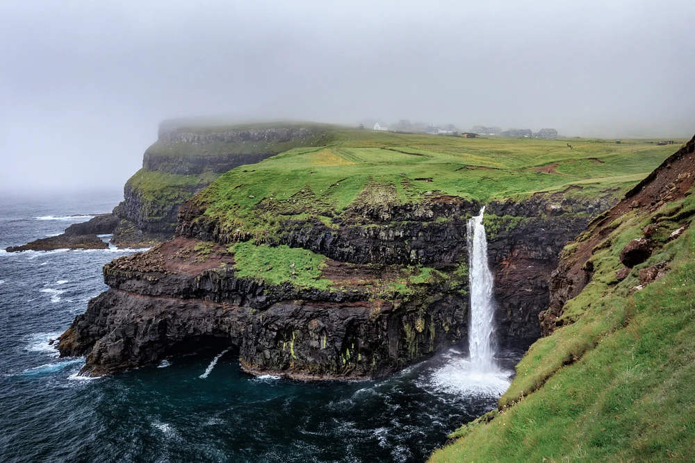 M&uacute;lafossur waterfall plunges from a green V&aacute;gar cliff into a misty Faroe Islands sea cove