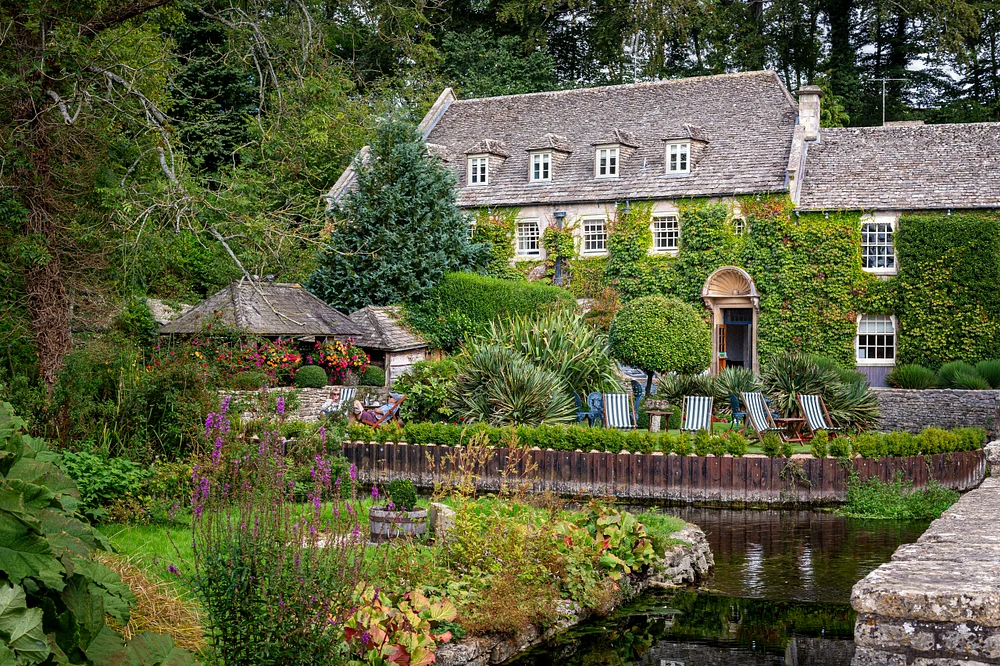 Ivy-covered Bibury cottage beside a flower-filled garden, stone stream channel, and leafy woodland in England