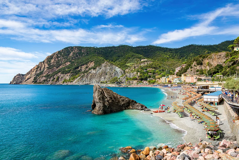 Turquoise Monterosso bay curves beneath wooded cliffs, colorful waterfront buildings, and a rocky Ligurian headland
