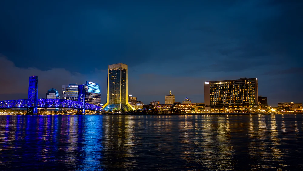 Night view of Jacksonville skyline with illuminated buildings and blue-lit bridge reflecting on the river.