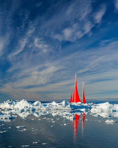 Vibrant red sailboat navigating through icy waters under a blue sky.