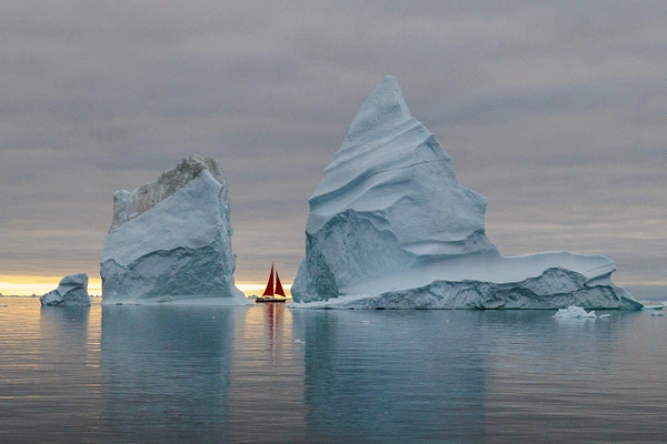 A red sailboat glides between towering icebergs in calm Arctic waters.