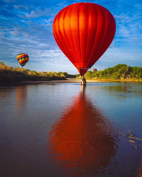 Two colorful hot air balloons floating above a calm river with reflections on the water.
