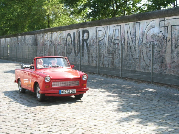 Red Trabant convertible driving past the Berlin Wall with graffiti art.
