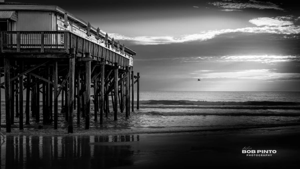 Black and white photograph of a wooden pier over ocean waves beneath a dramatic cloudy sunset sky